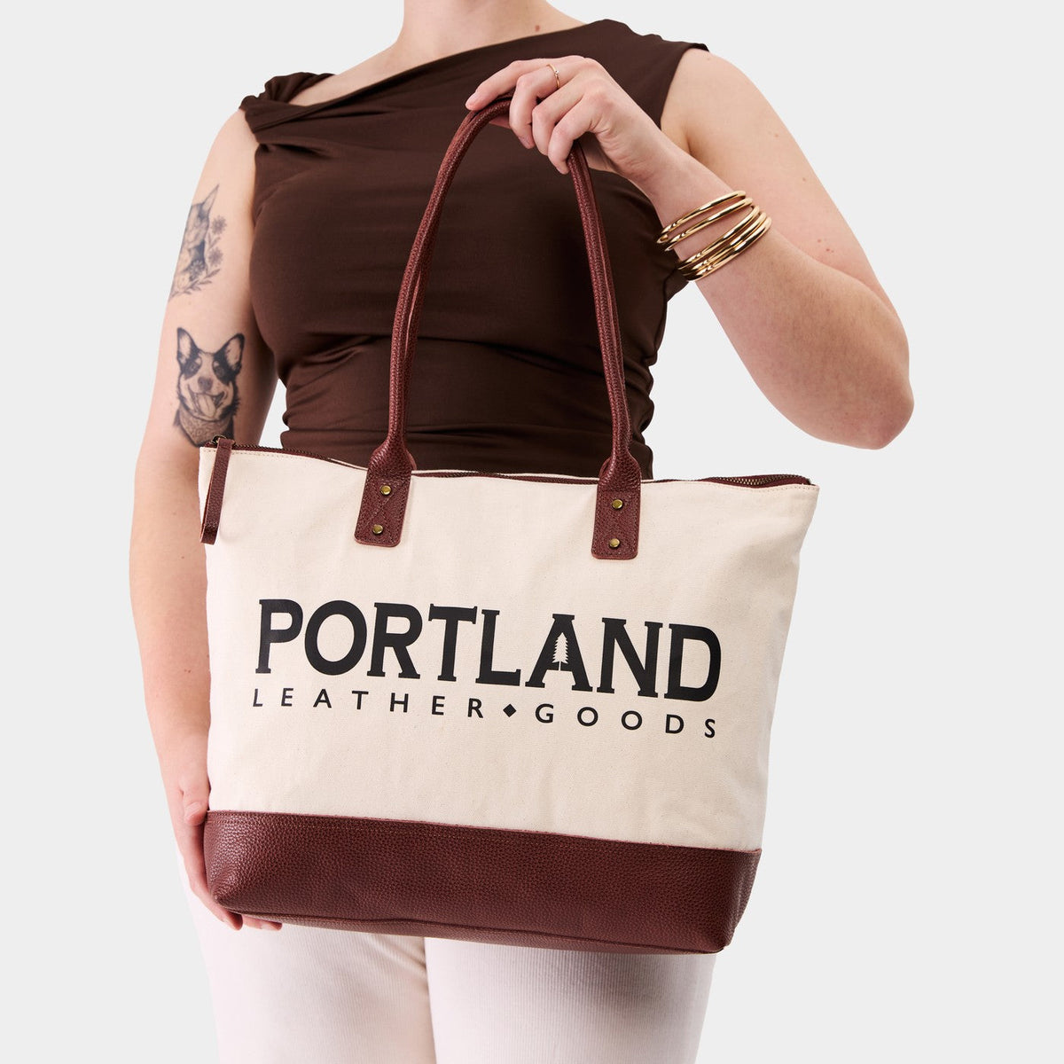 Model holding canvas tote with zipper, leather bottom, and leather handles on a white background
